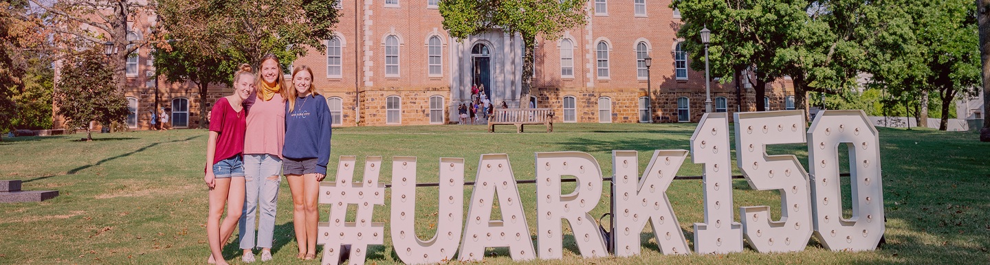 three students standing next to UARK150 sign
